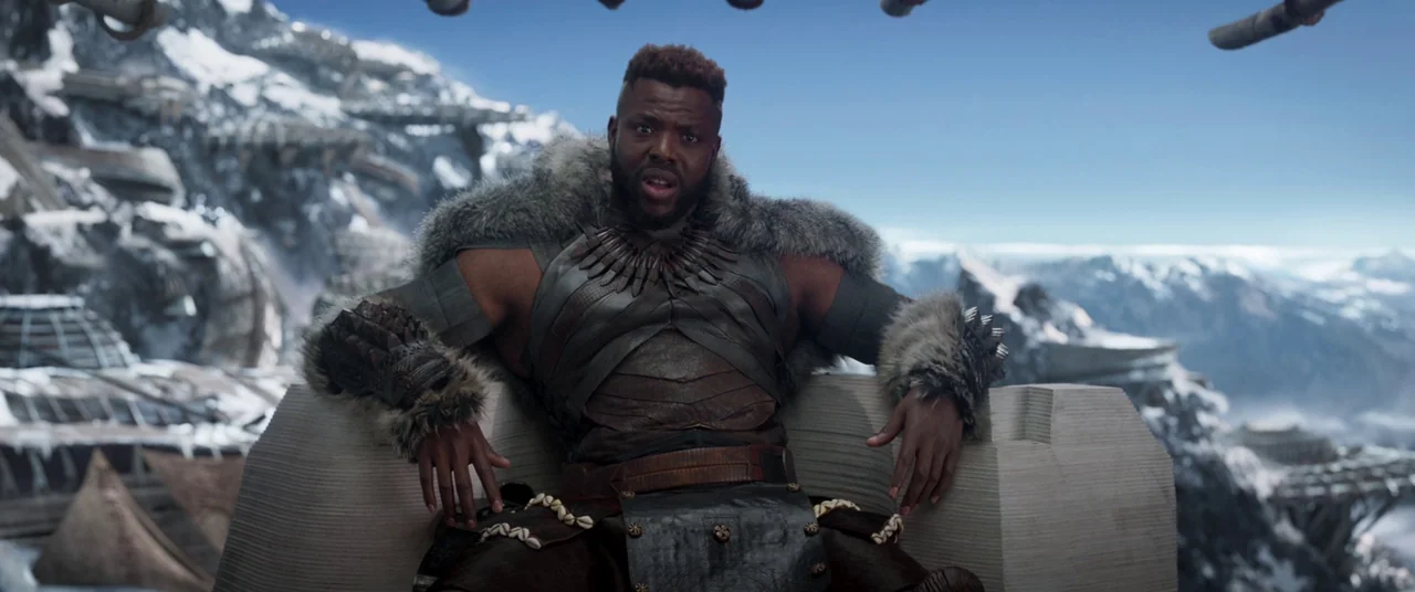 Black Panther (2018) movie still: medium — A man sits on a stone throne overlooking a high snowy mountain range. The background features futuristic…