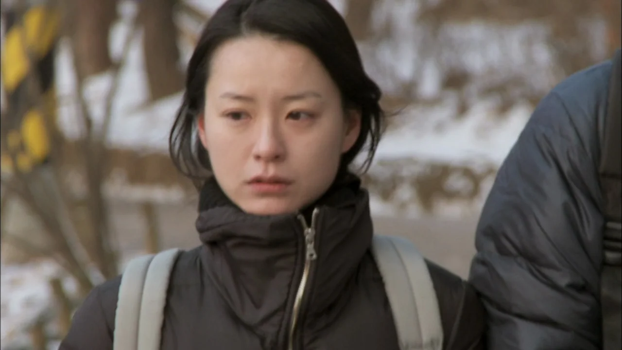 Oki’s Movie (2010) movie still: medium — A woman stands in a snowy park setting during the day. She wears a black puffer jacket and carries a…