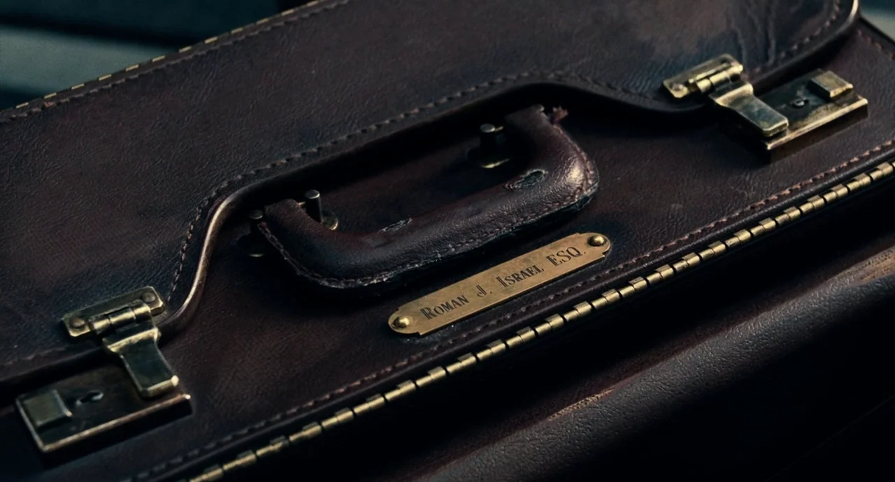 Roman J. Israel, Esq. (2017) movie still: closeup — A well-worn dark brown leather briefcase features a brass nameplate identifying the owner. The…