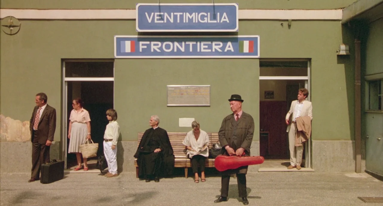 The Belly of an Architect (1987) movie still: wide — A green-painted station building with signs reading Ventimiglia Frontiera stands in bright…
