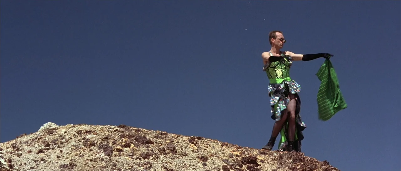 The Adventures of Priscilla, Queen of the Desert (1994) movie still: fullbody — A figure stands on a sunlit rocky incline under a clear deep blue…