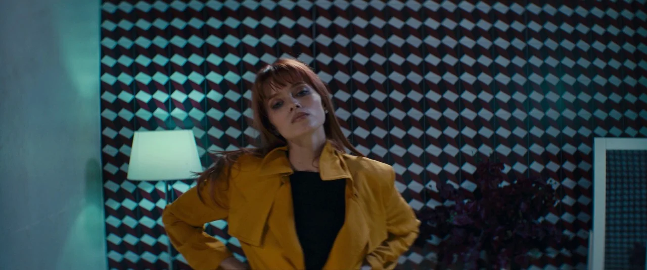 Elizabeth Harvest movie still: medium — A wall with a bold black and white geometric diamond pattern serves as backdrop. A woman in a yellow coat…