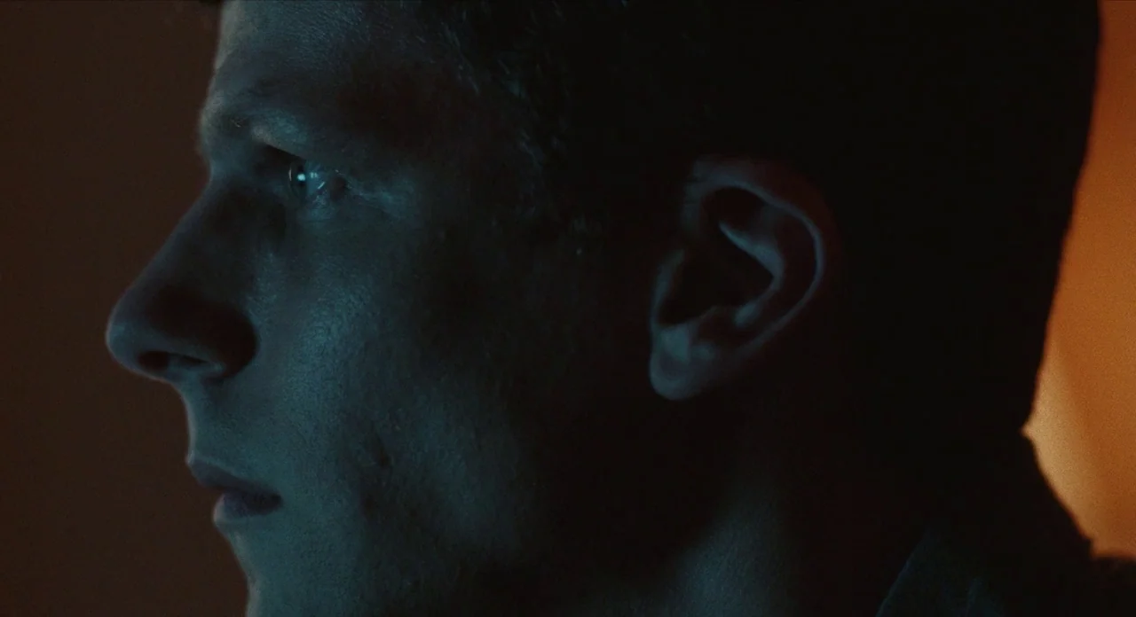 The Art of Self Defence (2019) movie still: closeup — A dimly lit indoor space with heavy shadow and minimal background detail. A man is viewed in…