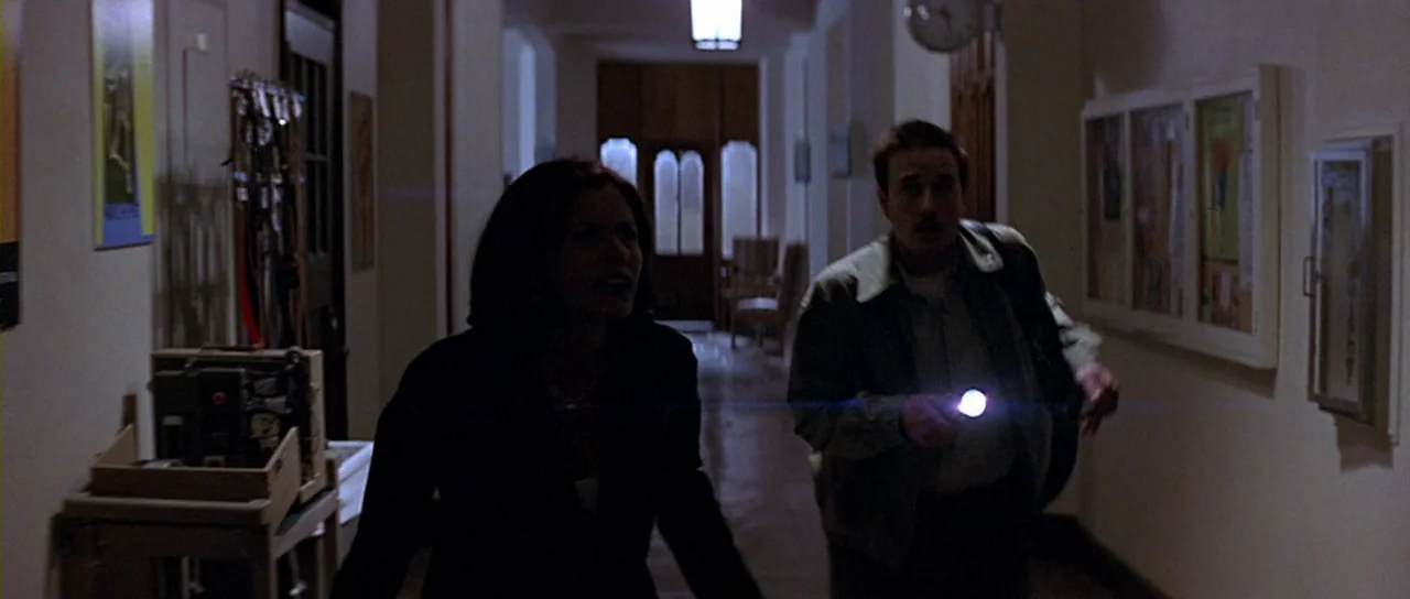 Scream 2 (1997) movie still: fullbody — A dimly lit indoor hallway with off-white walls and posters framed on the side. A woman and a man holding a…