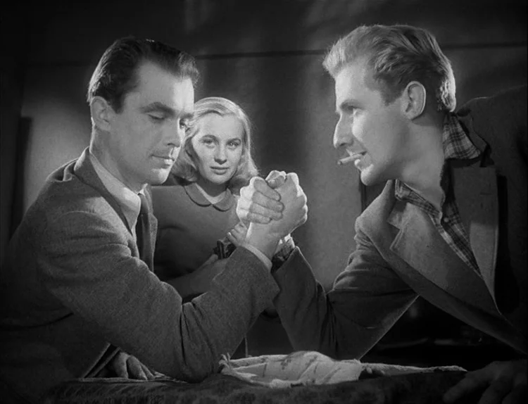 Music in Darkness (1948) movie still: medium — Two men arm-wrestle in a dark space while a woman watches in the background. The men are positioned…