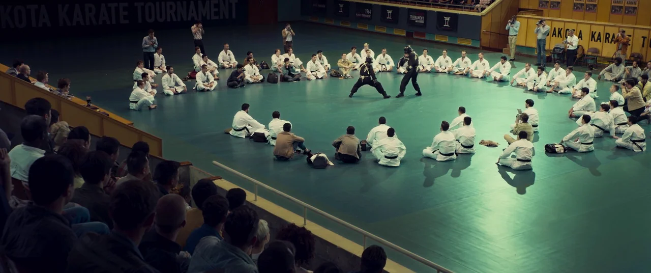 Ip Man 4: The Finale (2019) movie still: wide — A large indoor arena with a green mat floor and many spectators sitting around. Two practitioners in…