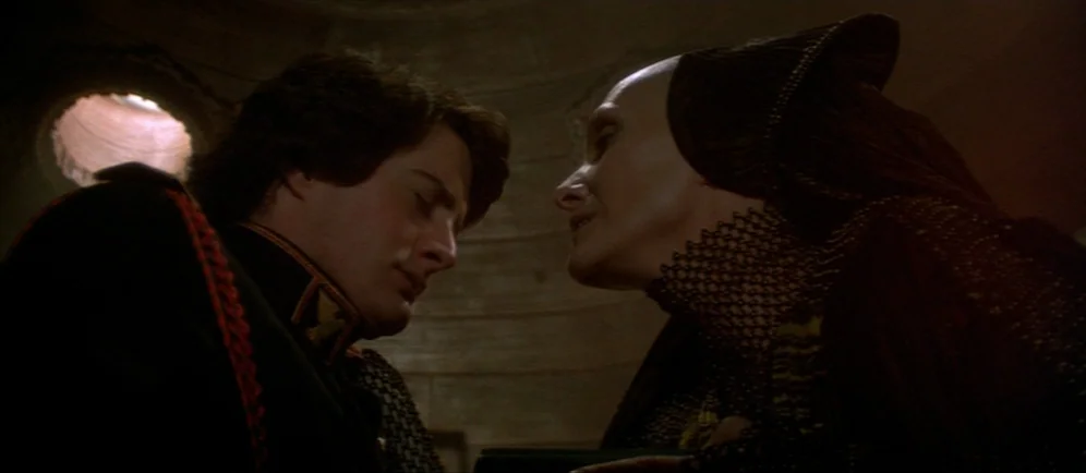 Dune (1984) movie still: medium — A dark stone chamber features a circular vent above the subjects. A young man stands opposite an elderly woman in…