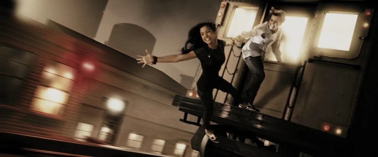 The Spirit (2008) movie still: fullbody — A woman balancing on the exterior side of a moving train car at night. She reaches toward a man who is…