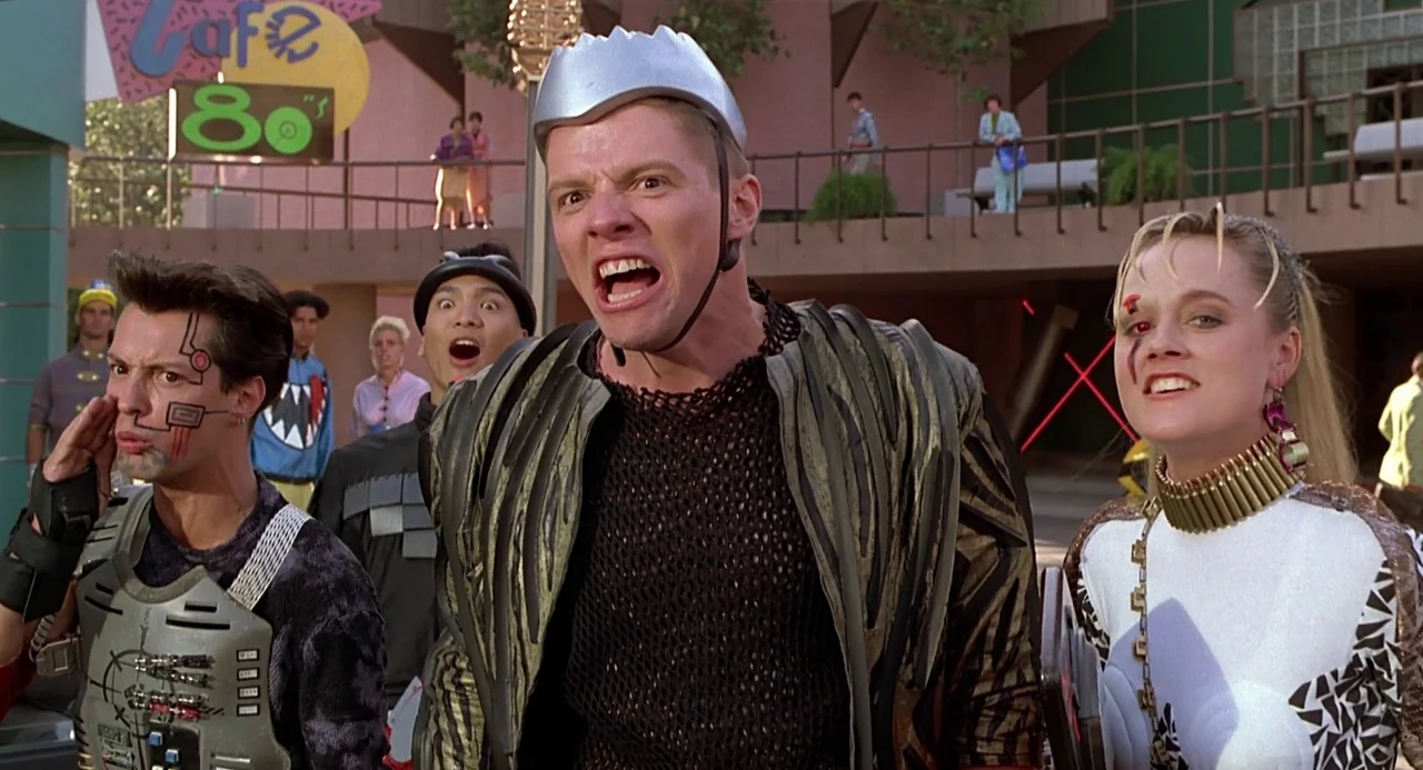 Back to the Future II (1989) movie still: medium — A bustling outdoor plaza near a shopping center features 1980s architecture and signage. A group…