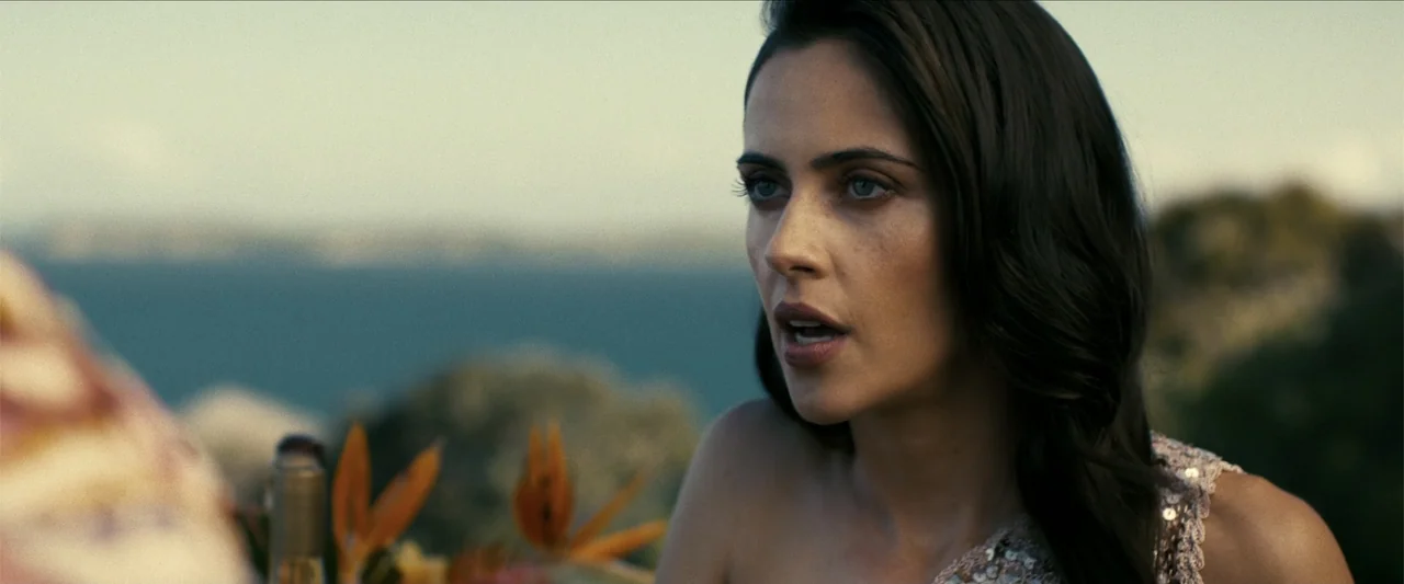 The Wrecking Crew (2026) movie still: closeup — An outdoor coastal area with blurred water and rock formations in the distance. A woman with dark…