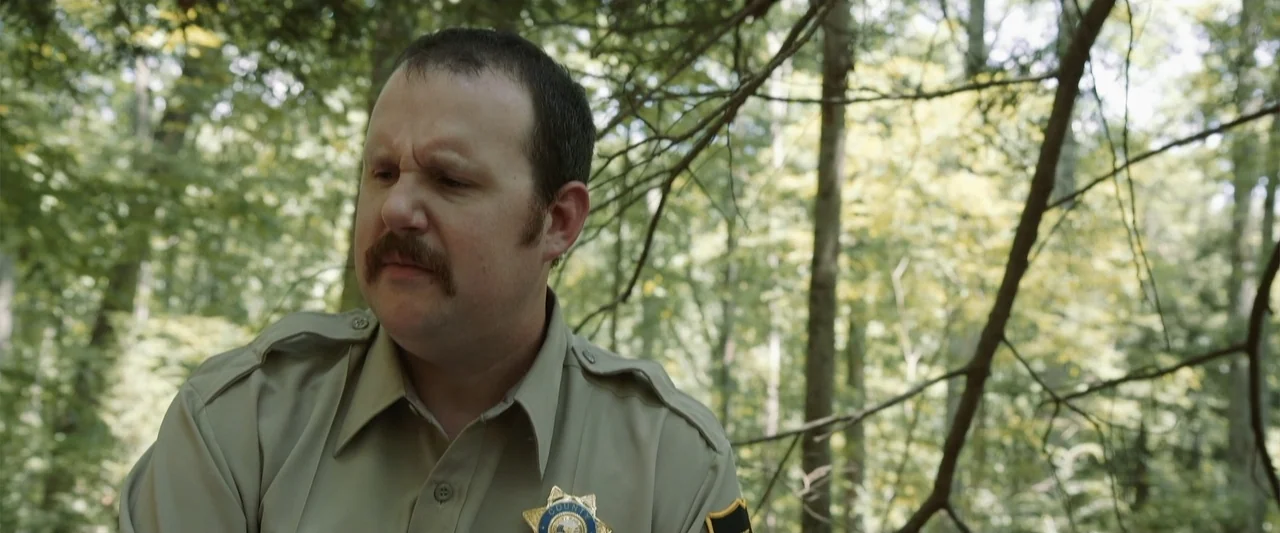Darkness Waits (2020) movie still: medium — A dense patch of woodland with mature trees fills the background behind the subject. A man in a law…