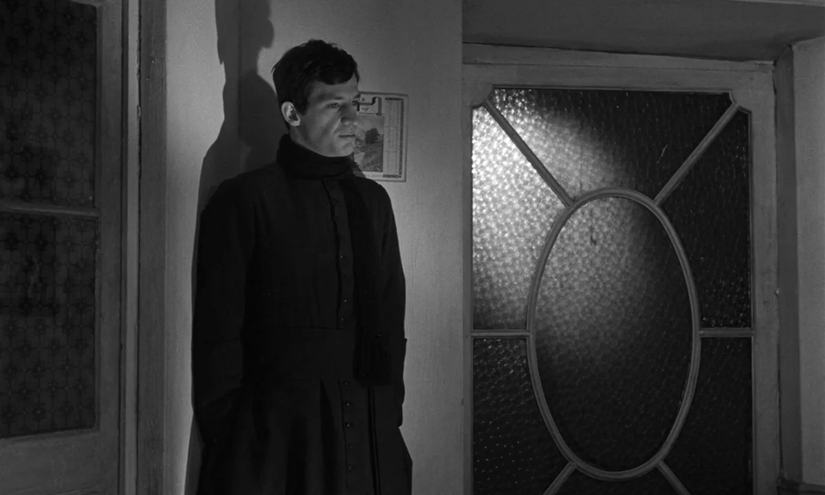 Léon Morin, Priest (1961) movie still: medium — A dimly lit indoor space featuring a door with textured glass panels. A man wearing a clerical…