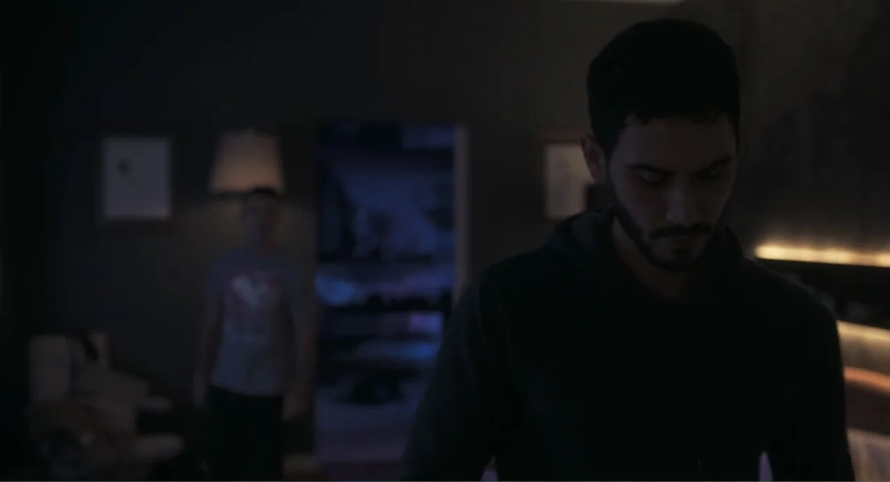 Straight (2024) movie still: medium — A dark bedroom with a lamp on a side table and a blurred doorway in the background. A man stands in the…