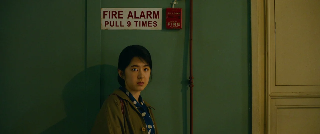 Swing Kids (2018) movie still: medium — A woman stands in a green hallway beneath a fire alarm instruction sign. She looks directly forward while…