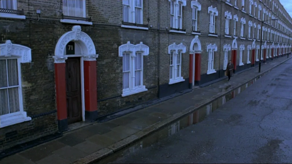 Spider (2002) movie still: wide — A long row of identical brick townhouses lines a wet city street at midday. A lone figure walks along the sidewalk…