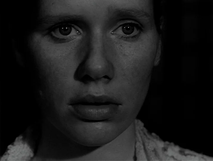 Hour of the Wolf (1968) movie still: closeup — A close-up view shows a woman's face emerging from a deep black void. Her eyes appear wide as she…