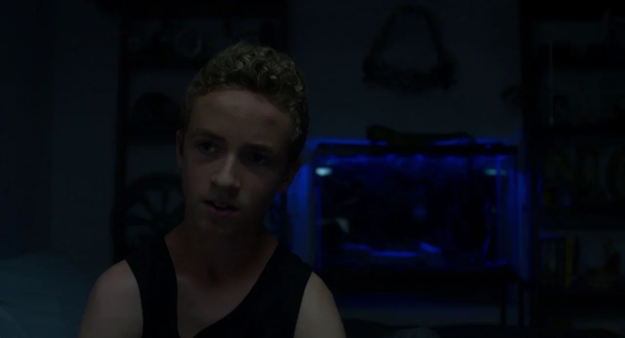 Maps to the Stars (2014) movie still: medium — A dark, dimly lit space contains a glowing blue fish tank in the background. A teenage boy sits in…