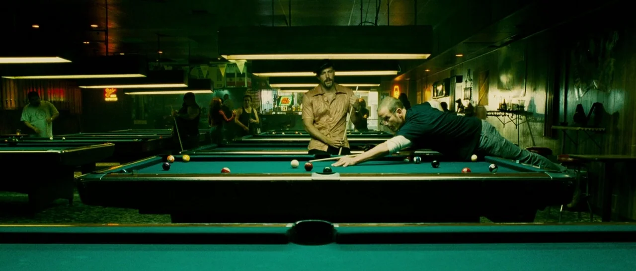 Slice (2018) movie still: wide — A dimly lit pool hall features several green felt tables under low-hanging rectangular lights. One man stretches…