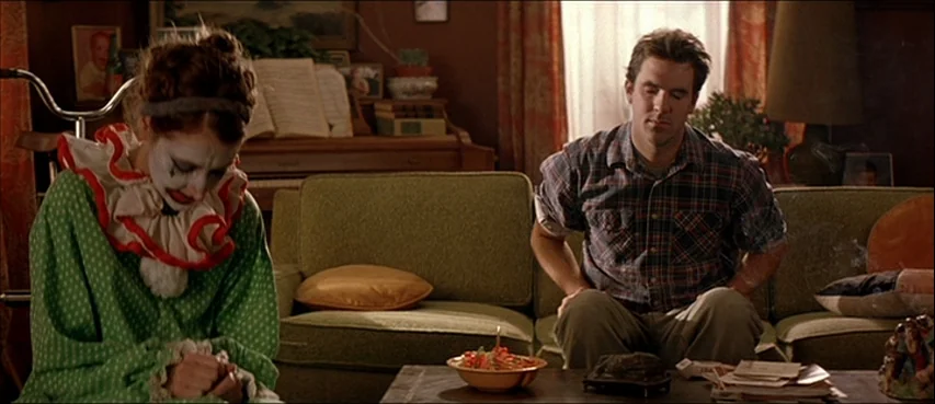 All The Real Girls (2003) movie still: medium — A cluttered suburban living room with a green sofa and an upright piano behind. A clown and a man in…
