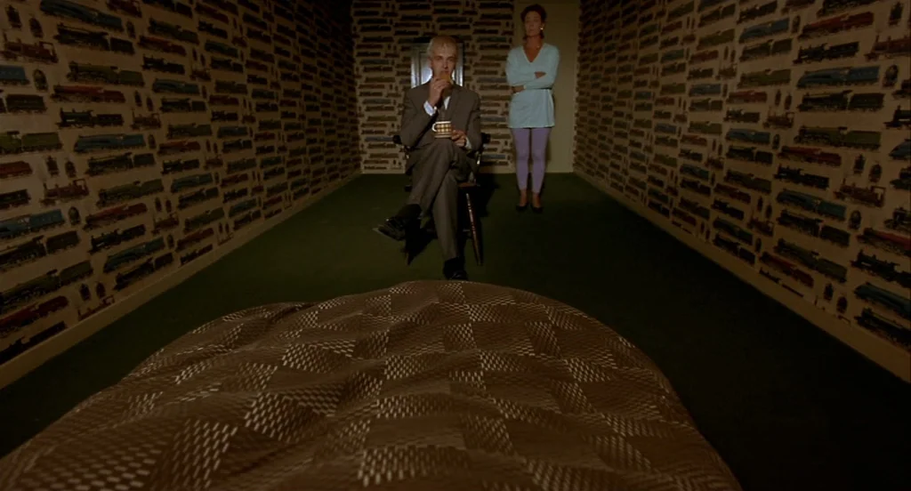 Trainspotting (1996) movie still: wide — A room with patterned wallpaper featuring toy trains and a dark green carpet floor. A man sits on a wooden…