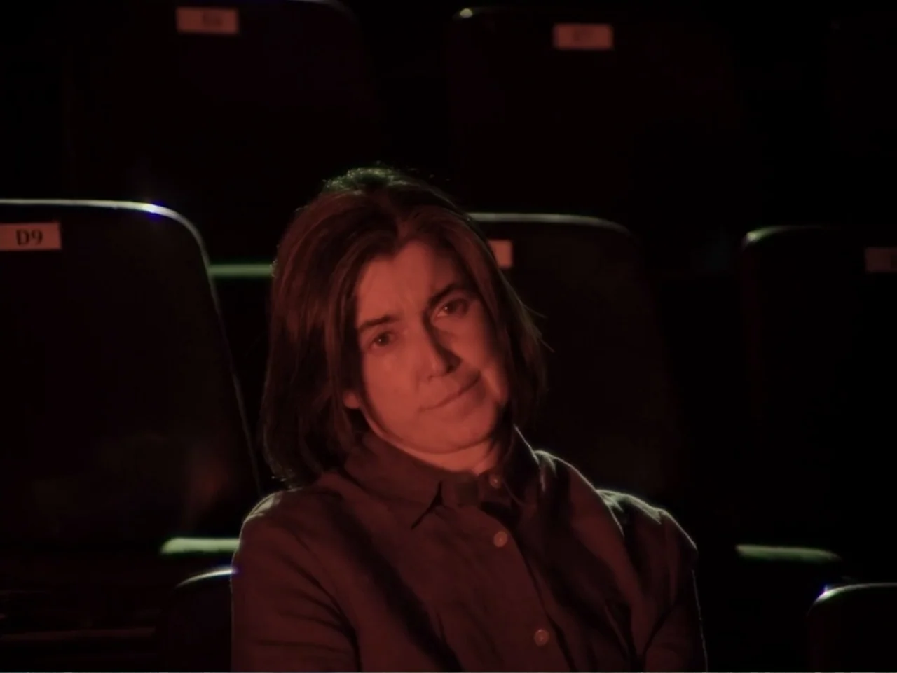 History and Geography (2024) movie still: medium — A dark movie theater interior with rows of empty seats visible in shadows. A woman sits alone in…