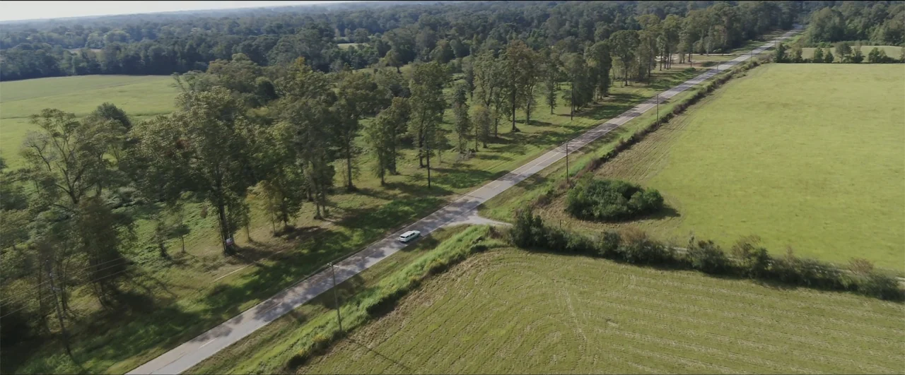 Jeepers Creepers: Reborn (2022) movie still: establishing — A rural highway stretches through a landscape of expansive green fields and mature…