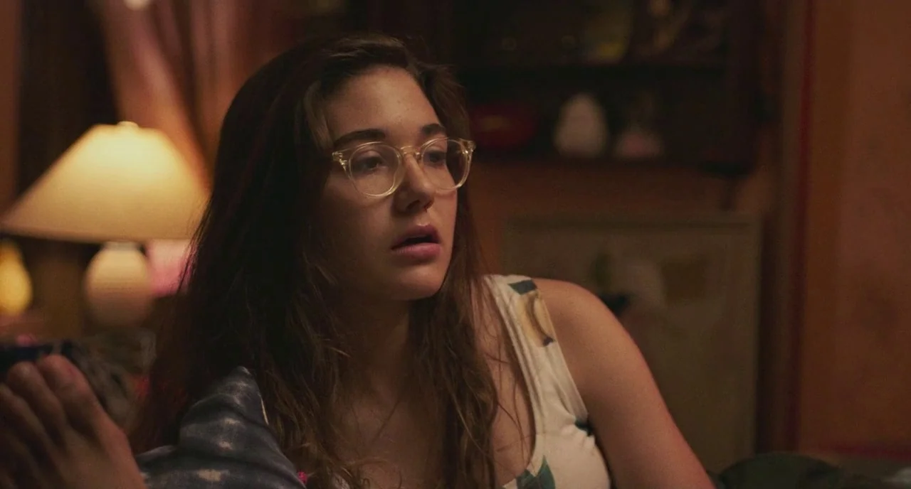 Skate Kitchen (2018) movie still: medium — A dimly lit room features a glowing table lamp against a shadowed background. A young woman with glasses…