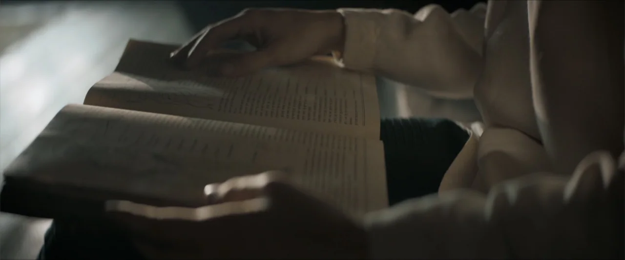 Dune (2021) movie still: closeup — A person reads an open book in a dimly lit interior room. The subject holds the book open while seated with hands…
