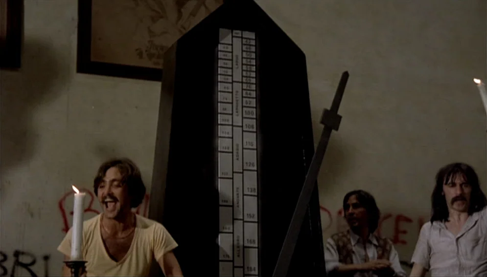 Orchestra Rehearsal (1978) movie still: medium — A dimly lit room features a giant black wooden metronome against a graffiti-stained wall. Three men…
