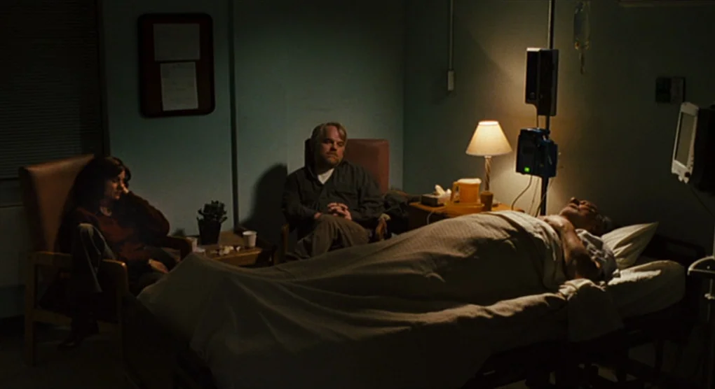 The Savages (2007) movie still: wide — A dimly lit room with blue walls and a central medical bed with a patient. A man and a woman sit in chairs…