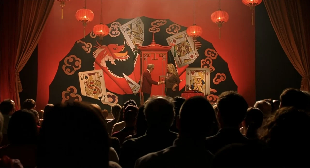 Scoop (2024) movie still: wide — A theatrical stage features a large red backdrop decorated with playing cards and a dragon. A man and a woman stand…