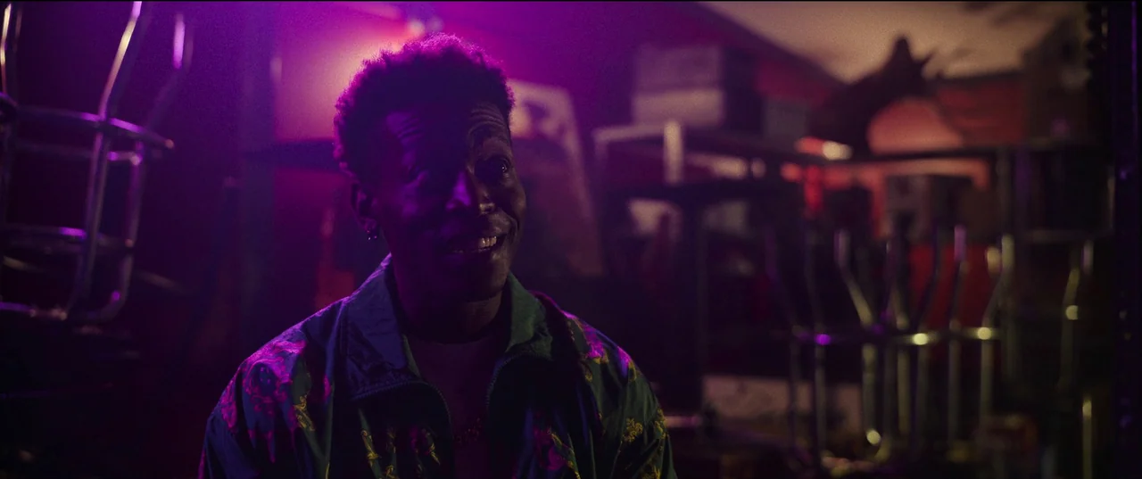 Hit Man (2023) movie still: medium — A dark, cluttered storage room is illuminated by vibrant, colored neon light sources. A young man wearing a…