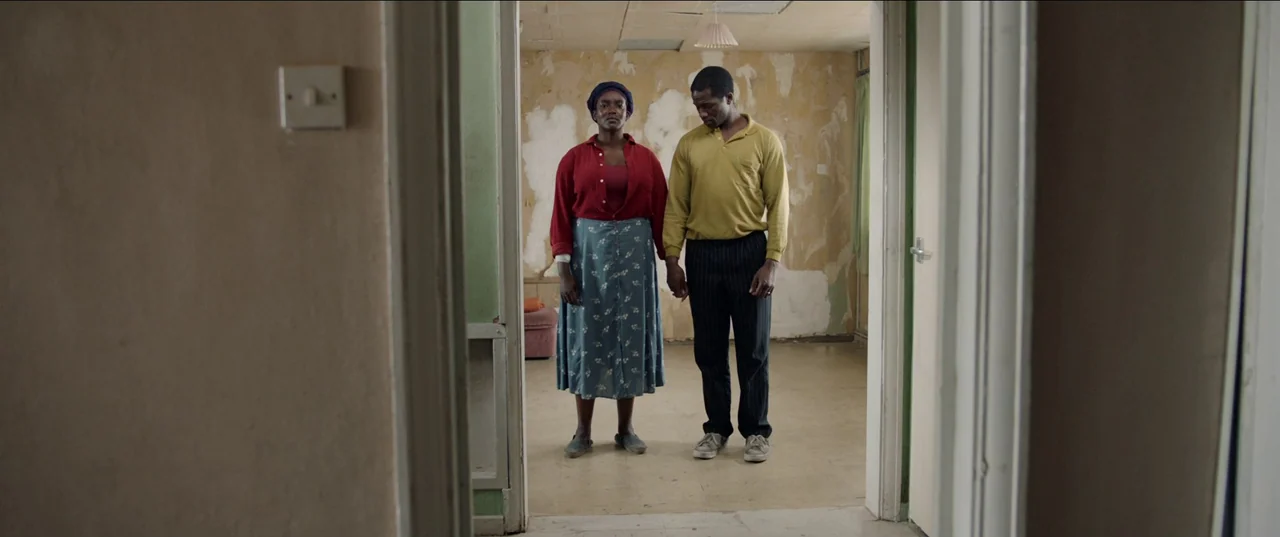 His House (2020) movie still: fullbody — A dilapidated room features walls with peeling beige wallpaper and exposed plaster patches. A man and a…