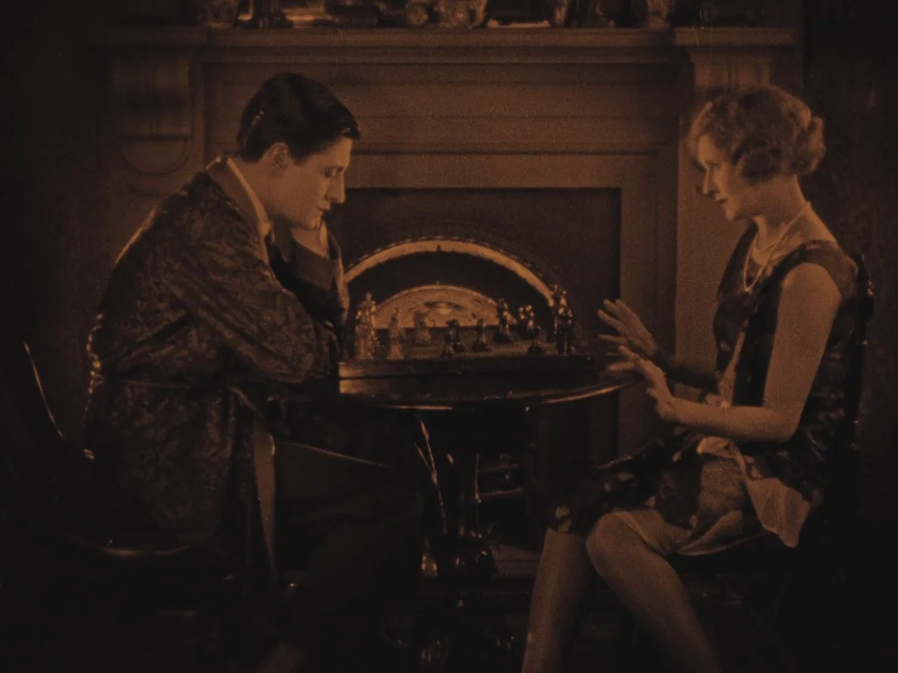 The Lodger (1927) movie still: medium — A dark living room features a fireplace mantel acting as the background for two people. A man and a woman…