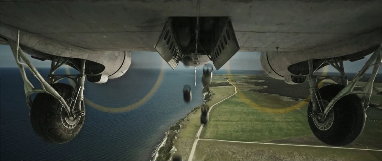 Sisu: Road to Revenge (2025) movie still: wide — A military aircraft flies over a coastal road running between sea and farmland. Multiple bombs fall…