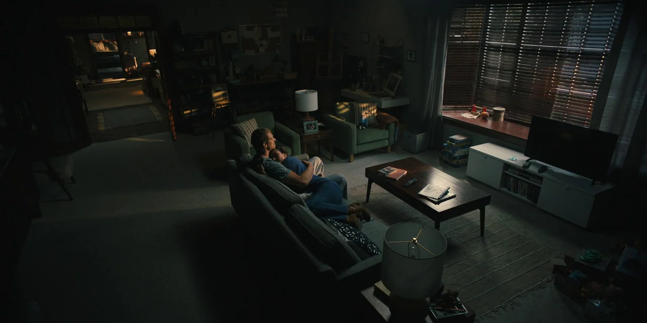 Silent Night (2023) movie still: wide — A dimly lit living room features a sofa, a coffee table, and a television. A man and a woman sit together on…