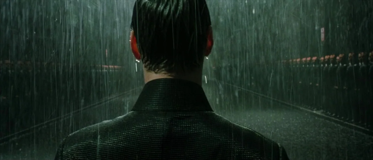 The Matrix Revolutions (2003) movie still: medium — A dark outdoor space is filled with heavy falling rain and long shadows. A man seen from the…