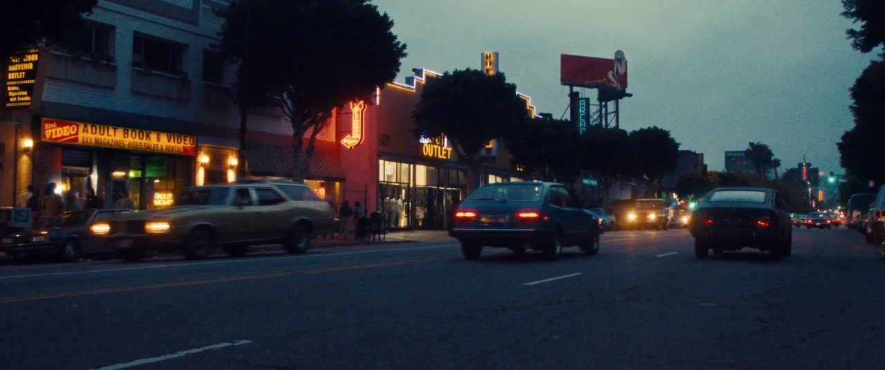 MaXXXine (2024) movie still: wide — A city street at twilight features low-rise buildings with neon signage for businesses. Several vintage cars…