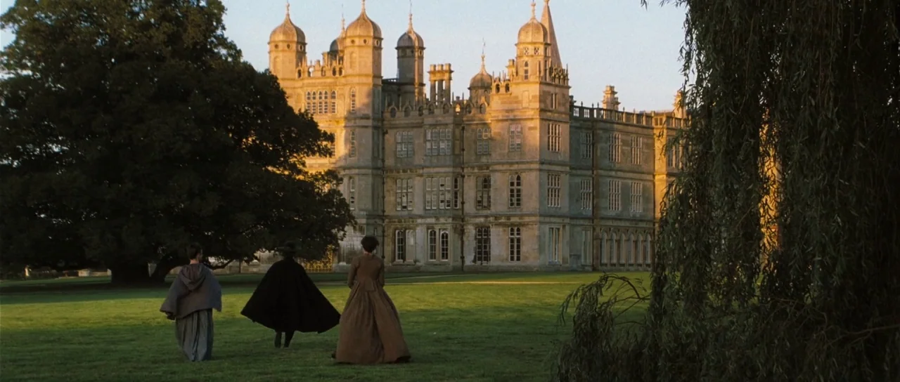 Pride & Prejudice (2005) movie still: wide — A sprawling stone manor stands behind three figures walking across an expansive manicured lawn. The…