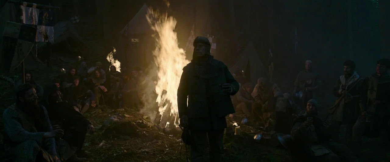 The Last Duel (2021) movie still: wide — A medieval military encampment is set in a dark forest at night. Soldiers sit around a tall fire pit near…
