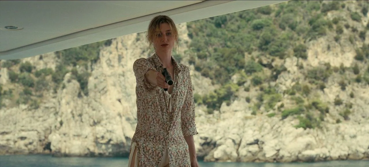 Tenet (2020) movie still: medium — The deck of a boat sits before a steep, pale, rocky cliffside near water. A woman wearing a floral shirt holds a…