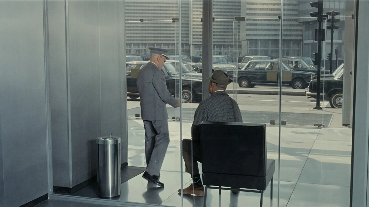 Playtime (1967) movie still: fullbody — A glass-walled modern lobby overlooks a busy city street with multiple parked cars. An official in a uniform…