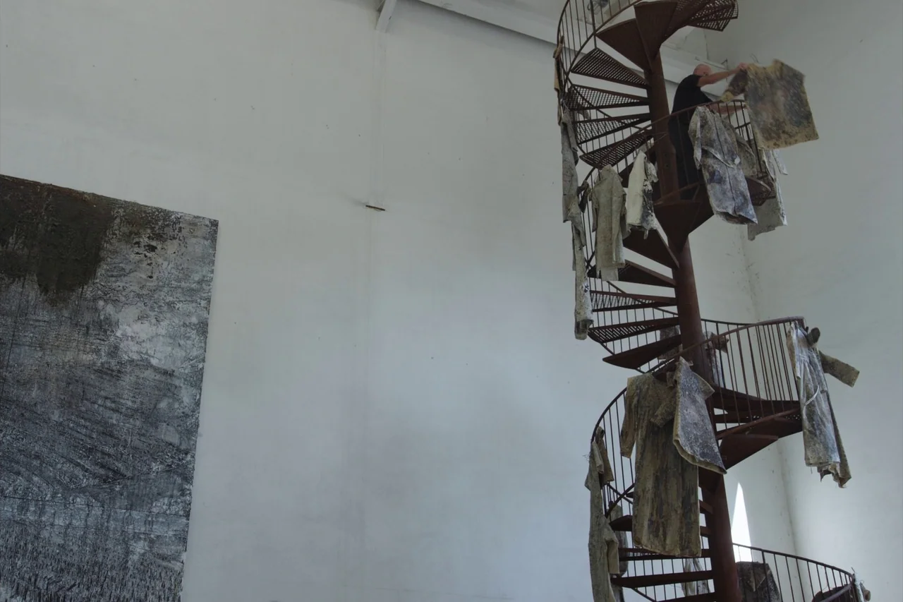 Anselm (2023) movie still: wide — A spacious white-walled exhibition area contains a large metal spiral staircase and wall art. A bald man climbs…