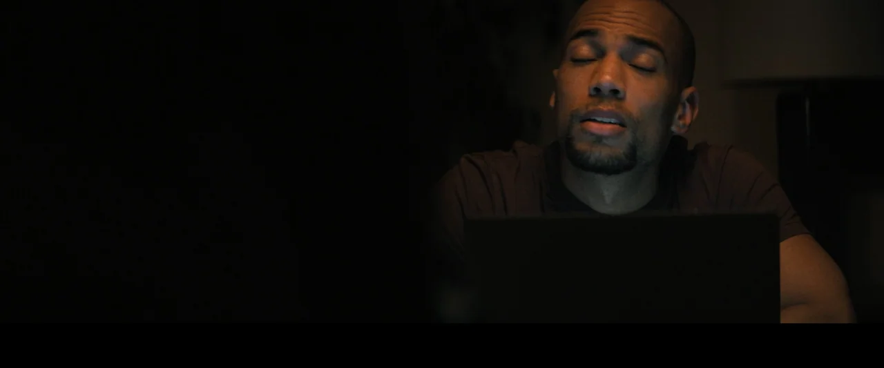 Something from Tiffany's (2022) movie still: medium — An interior space lit primarily by the glow from a computer screen at night. A man sits at a…