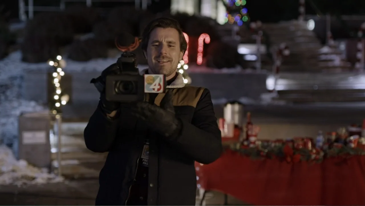 Peppermint and Postcards (2023) movie still: medium — An outdoor evening setting features holiday lights and a table with supplies nearby. A man…