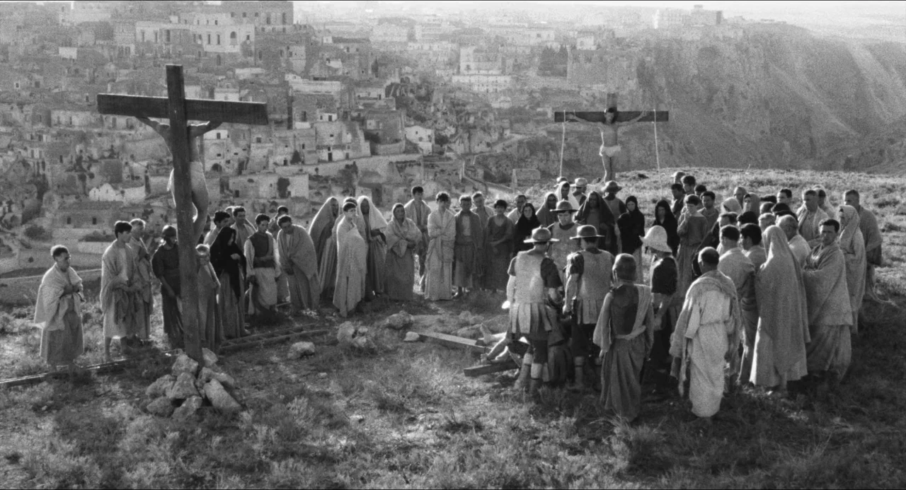 The Gospel According To St. Matthew (1964) movie still: wide — A crowd gathers on a rocky, grassy hilltop overlooking a distant ancient stone city.…