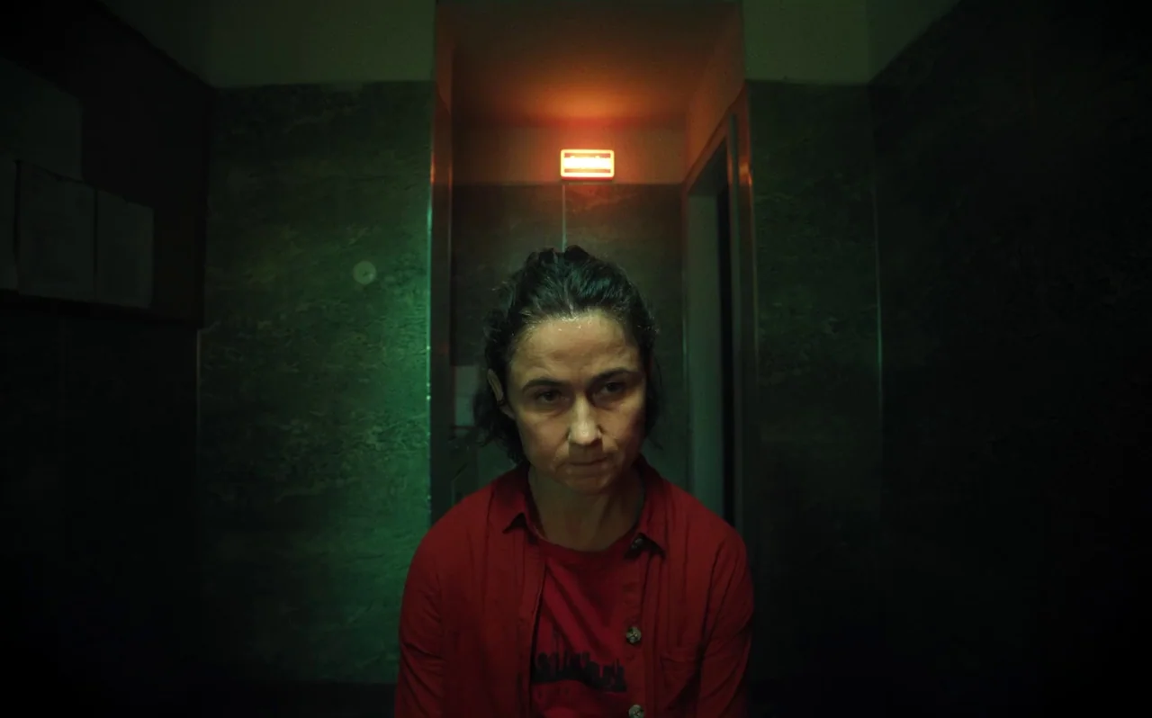 Knocking (2021) movie still: medium — A dimly lit concrete stairwell with dark marble-like wall panels. A lone woman sits in the center, bathed in…