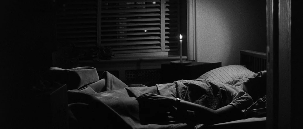 Manhattan (1979) movie still: medium — A dark, spare bedroom features a single window with closed blinds and a bedside table. A lone person lies…