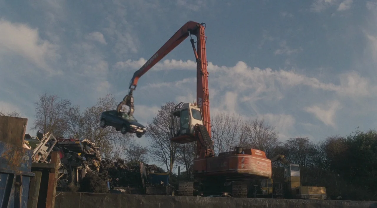 Broken (2012) movie still: wide — An industrial scrap yard features a large orange excavator operating under a cloudy sky. The heavy equipment crane…