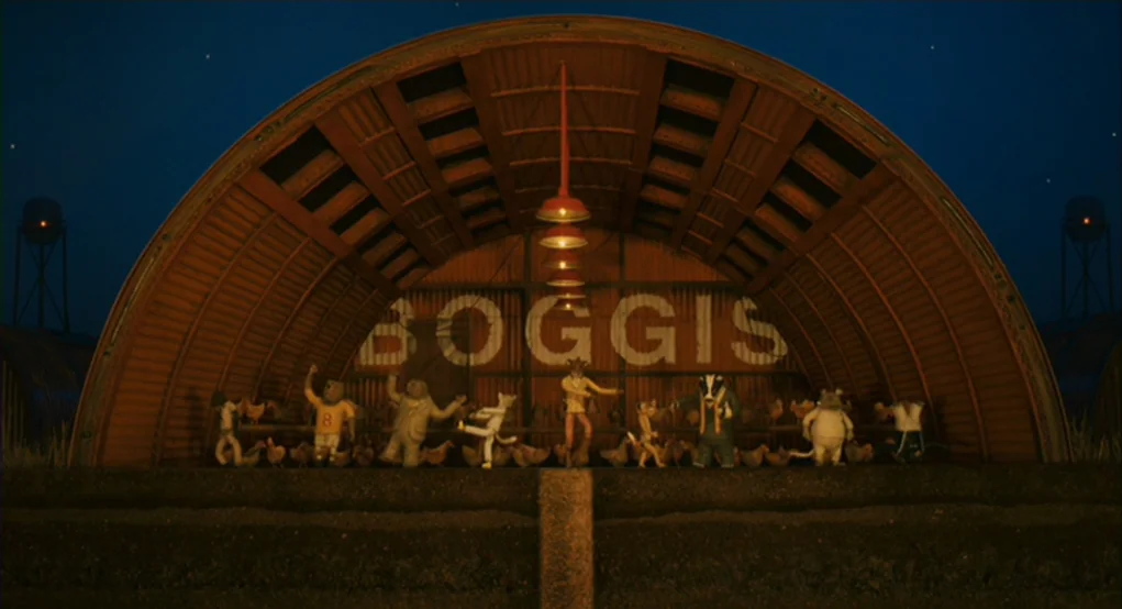 Fantastic Mr. Fox (2009) movie still: wide — An arched corrugated metal structure with a large BOGGIS sign occupies the background. A group of…
